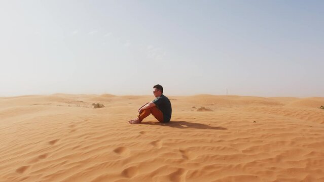 Young barefoot man sits on sand in desert and looks into smartphone. concept of solitude and reflection. Slow motion