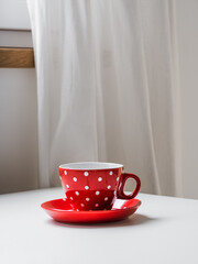 A red coffee cup with white polka dots stands on a white table. White linen curtain in the background. Bright kitchen in the morning. minimalism. Place for text. copy space. Vertical