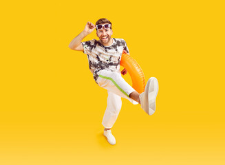 Fototapeta na wymiar Happy guy enjoying summer holidays at the sea. Cheerful man in trendy clothes holding an inflatable beach ring walking and dancing isolated on a yellow orange color background. Full length portrait