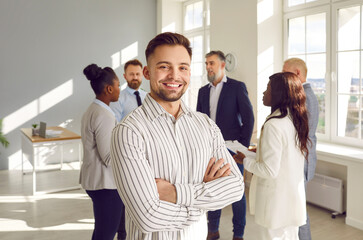 Portrait of a young happy smiling confident bearded business man leader proudly looking at the camera with crossed arms with team of company employees talking in background in the office.