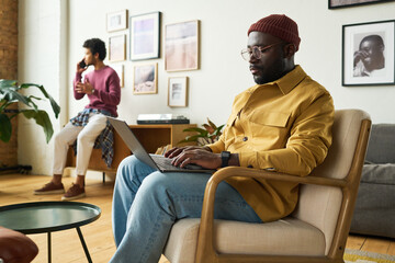 Young serious African American analyst typing on laptop keyboard while sitting in armchair against male coworker talking on smartphone