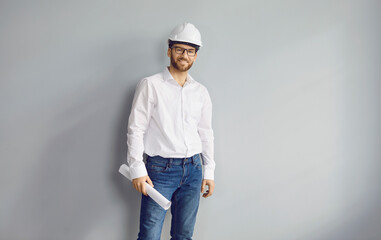 Happy smiling adult man architect wearing helmet and holding blueprint isolated on grey background. Construction engineer in glasses wearing jeans and white shirt looking cheerful at the camera. - Powered by Adobe