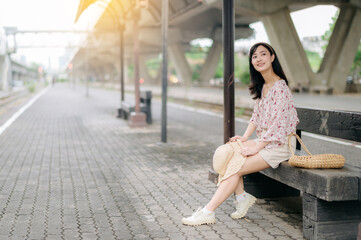 Fototapeta premium young asian woman traveler with weaving basket waiting for train in train station. Journey trip lifestyle, world travel explorer or Asia summer tourism concept.