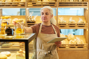 Aged female owner of bakery shop with tablet in hand standing by glass display with fresh pastry and looking at jar containing cookies