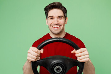 Young smiling happy fun cheerful satisfied man he wears red t-shirt casual clothes hold steering wheel driving car isolated on plain pastel light green background studio portrait. Lifestyle concept.