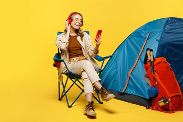 Full body young woman sit near bag stuff tent use mobile cell phone listening music in headphpnes...