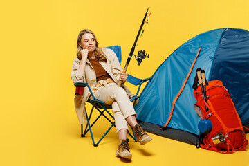 Full body minded young woman sit near bag with stuff tent hold fishing rod isolated on plain yellow background. Tourist leads active lifestyle walk on spare time. Hiking trek rest travel trip concept.