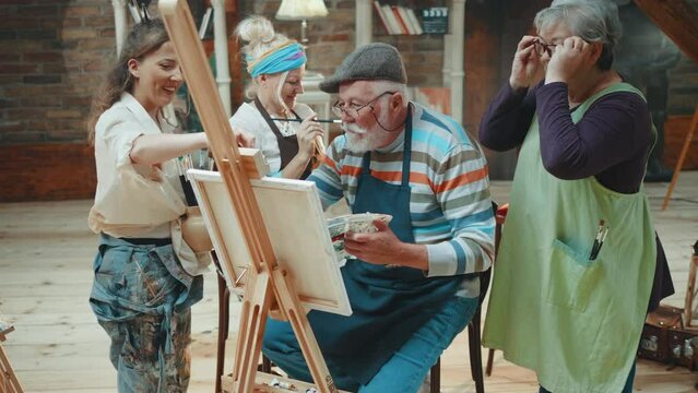 An old man paints on a canvas while colleagues watch him having a group art class