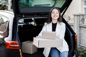 The girl sits in the trunk of the car and shows the boxes forward in front of the camera. Concept of buying goods online and delivering them home