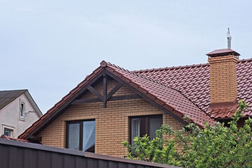 brown brick attic of a private house with windows under a tiled roof with a chimney behind a fence on the street against a gray sky