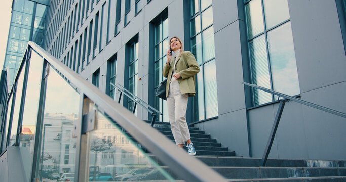 Attractive young caucasian woman walking down the stairs speaking on mobile phone background of a modern business center. Girl talking friendly conversation smiling cheerfully outdoor.