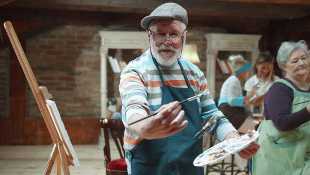 Happy old man with painting brush in hand smiling and looking at camera while standing in art studio at group painting class