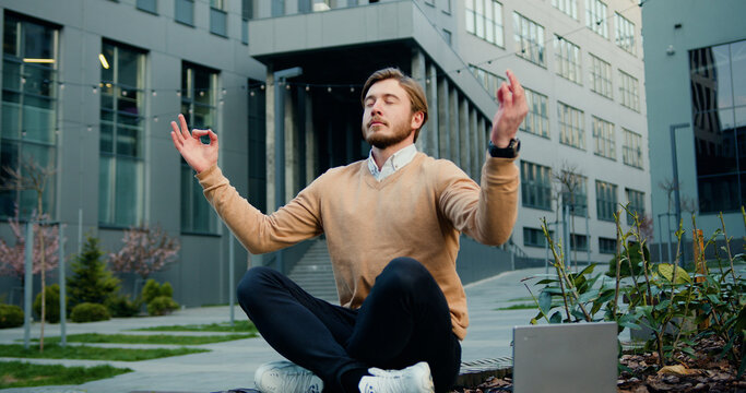 Handsome office man relaxing in the lotus position, sitting in front of modern office building. Office workers doing yoga.