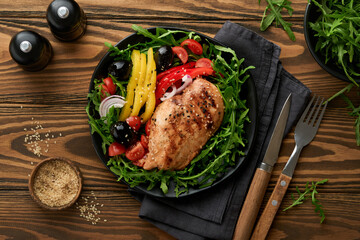 Salad with grilled chicken breas fillet with fresh arugula, tomatoes and olives on black plate on old wooden dark background. Chicken meat with salad. Ketogenic lunch. Healthy food. Top view.
