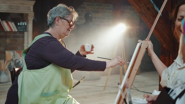 An elderly retired woman drinks coffee and paints on canvas while talking with colleagues in an art class