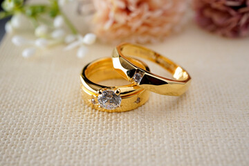 Couple gold wedding rings on a burlap cloth with brown flowers and cream on the back	