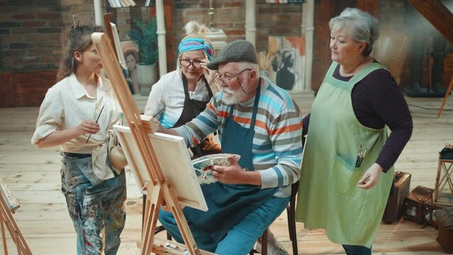 A group of retired artists observing an old male colleague painting on a canvas during a group painting class in a community center with a female teacher