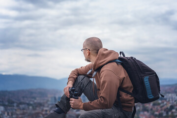 Young adult male traveler with backpack sitting on top overlooking the city Sarajevo with binoculars. travel to Balkans