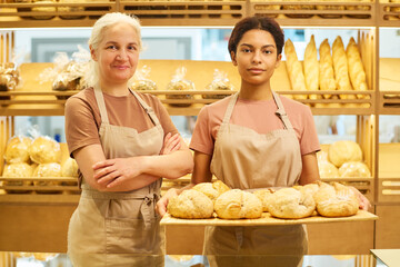 Two intercultural female bakers or clerks of bakery shop in uniform looking at camera while...