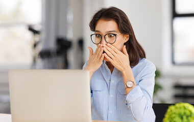 Shocked   woman freelancer looks in amazement at laptop screen and covers her mouth with hands in...
