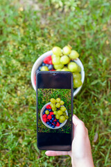 Miseczka wypełniona owocami maliny, borówki i winogrona, fotografowana za pomocą  telefonu komórkowego.