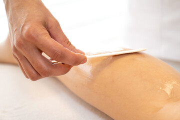 Close up of beautician's hand applying cooling gel with wooden stick on client's leg before laser hair removal treatment