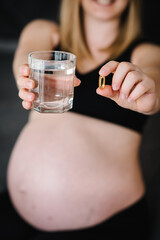 Pregnant woman take vitamins daily Omega 3 and glass water. Female hands hold supplements or vitamin. Girl sitting in bed at home. Pregnancy, treatment, medicine healthcare concepts. Photo of pills.