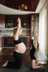 Pregnant woman and child in sport clothing exercising at home. Female with big belly. Family yoga concept. Pregnancy yoga and fitness. Mother and daughter training, meditating together, stands on mat.