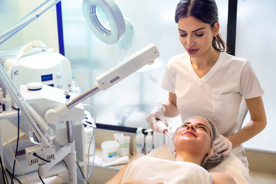 Female beautician using cosmetology electrical too to apply darsonval facial massage to beautiful young woman at beauty clinic.