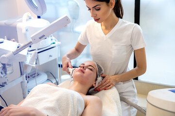 Beautician applying face treatment with modern apparatus on the face of young woman at the spa salon