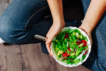 Green vegan breakfast meal in bowl with various fresh mix salad leaves and tomato. Girl in jeans sits on the floor and holding fork with knees and hands visible, top view on wooden background