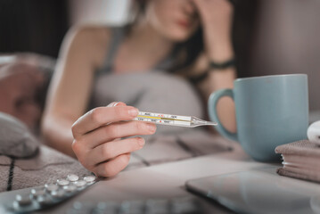 A young woman has a cold and lies in bed measures the temperature with a mercury glass thermometer closeup
