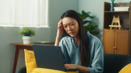 Serious Thoughtful Young Asian woman sit on sofa working at home living room. Asian female remote work using laptop computer looking at screen and thinking about problem solution or new business ideas