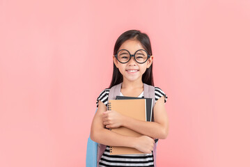 schoolgirl hugging book wearing backpack smiling isolated on pink background