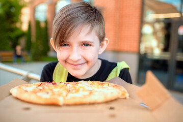 the boy holds  pizza, is going to eat. the child ordered pizza, sits on a bench with pizza in a box on a sunny day in the backyard of the school