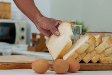 Close-up dining table in kitchen with fresh bread and eggs, An Asian man's hand reached in picked up several pieces bread but picked up just one piece, eat snacks as food in morning.