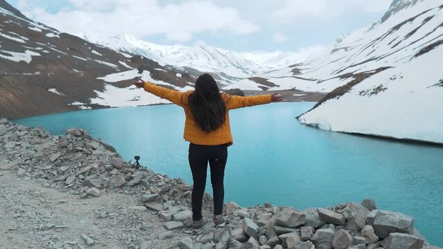 Rear view of Indian female enjoying snowfall with open arms at Suraj Tal Lake, Lahaul, India. Beautiful turquoise color water in Suraj Tal lake and snowy mountains during summer. Travel and holiday 