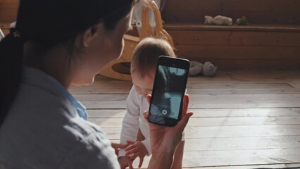 Over the shoulder shot of woman sitting on floor at home and making video of her barefoot baby making first steps with smartphone