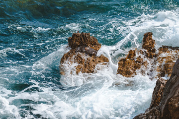 Ocean waves braking at coastline rocks covering them in white sea foam on sunny summer day