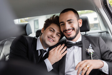 Cheerful homosexual groom with braces in suit hugging bearded boyfriend and looking at camera after...