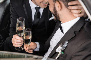 Smiling homosexual groom in formal wear holding champagne glass and hugging bearded boyfriend while...