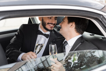 Positive and bearded homosexual groom in elegant suit holding glass of champagne and looking at young boyfriend while sitting on backseat of car during honeymoon