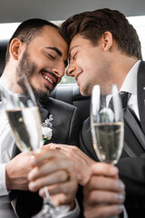 Portrait of overjoyed gay newlyweds in classic attire holding blurred hands and glasses of champagne during honeymoon road trip in car