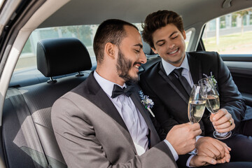 Smiling same sex grooms in formal wear toasting with glasses of champagne and holding hands while...