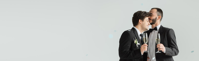 Young homosexual man in suit holding champagne and kissing smiling boyfriend with champagne under confetti during wedding ceremony on grey background, banner