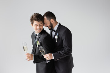 Smiling and bearded gay groom in elegant formal wear with boutonniere holding glass of champagne near boyfriend in braces with closed eyes during wedding celebration isolated on grey