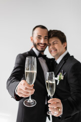 Blurred lgbt couple in elegant formal wear smiling and showing champagne glasses at camera during wedding celebration isolated on grey