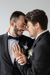 Portrait of young gay man with braces in elegant suit with boutonniere touching positive and bearded boyfriend during wedding ceremony isolated on grey