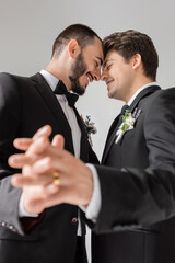Low angle view of cheerful gay couple in formal wear with floral boutonnieres holding blurred hands during wedding isolated on grey