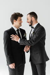 Cheerful and bearded gay man in elegant formal wear with floral boutonniere holding hand of groom during wedding celebration isolated on grey